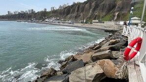 A rocky coastline with a lifebuoy, a pier, and buildings in the background.