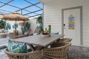 An outdoor patio with a wooden table, wicker chairs, a thatched umbrella, and a fruit bowl.