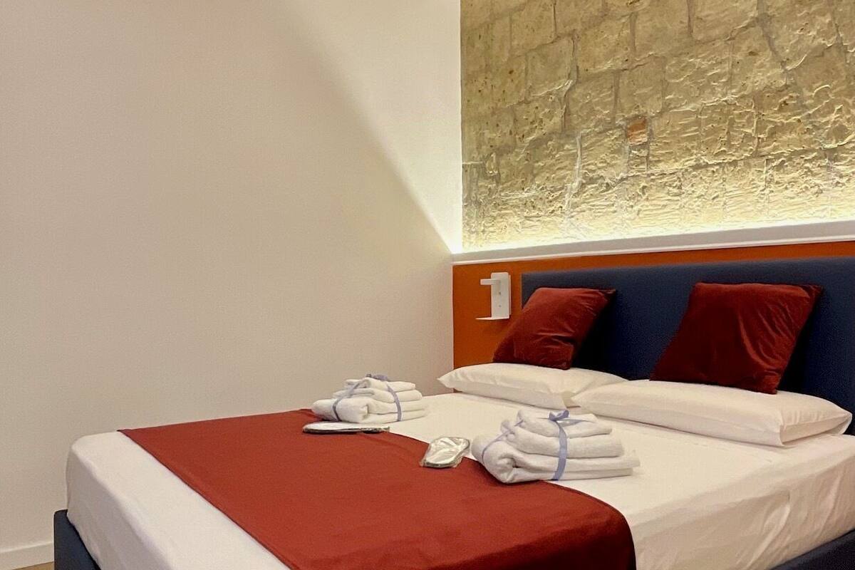 Une chambre d’hôtel avec un lit, une literie rouge et blanche, une tête de lit en bois et un mur en pierre.