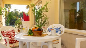 Una pequeña mesa redonda con una cesta de fruta y una taza, rodeada de sillas de plástico blanco con cojines estampados, en un balcón con plantas en macetas y vistas a un edificio con toldo rojo.