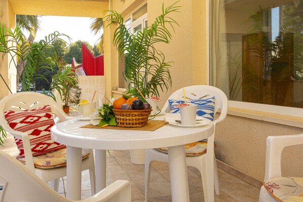 A small round table with a basket of fruit and a cup, surrounded by white plastic chairs with patterned cushions, on a balcony with potted plants and a view of a building with a red awning.
