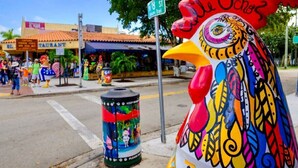 Una escultura de gallo colorido en una esquina de calle con un bote de basura que presenta un personaje de caricatura.