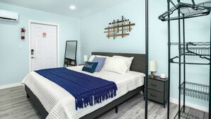 Un dormitorio ordenado con cama, mesita de noche, espejo en la pared, perchero y una puerta con extintor.