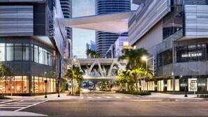 Una calle urbana moderna con edificios altos, un paso elevado peatonal y tiendas, incluyendo una tienda Cole Haan.