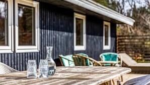 A rustic wooden table with three clear glasses and a bottle, set against a dark wooden building with white-framed windows. - Gorgeous home in Vejby with WiFi (Vejby)