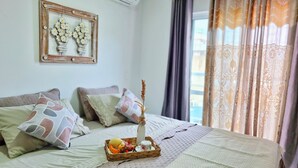 A neatly made bed with decorative pillows, a small table with a fruit basket and a candle, and a framed picture on the wall.