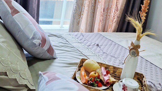 A neatly made bed with a tray of breakfast items, a vase with dried flowers, and a view of the outdoors through curtains.