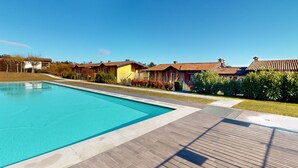 Une piscine entourée d’une terrasse en bois, avec des maisons aux toits rouges et un ciel d’un bleu limpide.