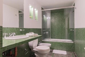 Un baño con pared de azulejos verdes, un lavamanos blanco, un inodoro y una ducha con mampara de vidrio.