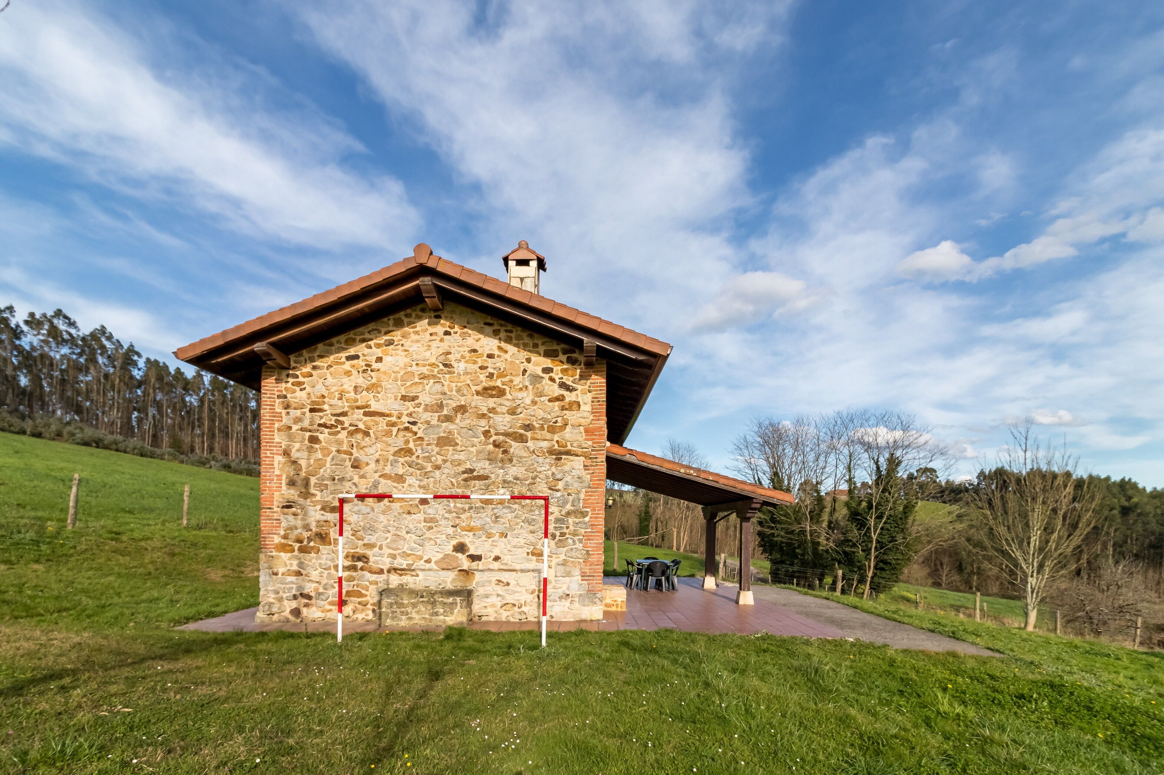 A stone building with a red roof, a covered patio, and a grassy area with trees in the background.