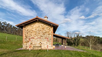 A stone building with a red roof, a covered patio, and a grassy area with trees in the background.