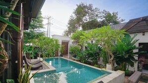 A rectangular swimming pool with a wooden deck, surrounded by lush tropical plants and a small building in the background. - Moin Villa Lembongan (Bali)