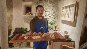 A person holding a wooden board with various meats and cheeses in a rustic kitchen with stone walls and a window.
