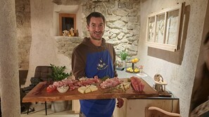 A person holding a wooden board with various meats and cheeses in a rustic kitchen with stone walls and a window. - Apartment 'Lauterfresser' with Mountain View, Shared Garden and Wi-Fi (Rodeneck)