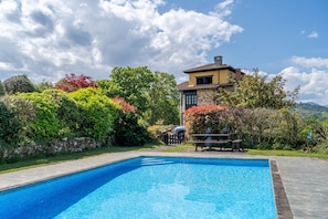 A swimming pool with a stone border, a stone wall, and a large house with a chimney in the background.