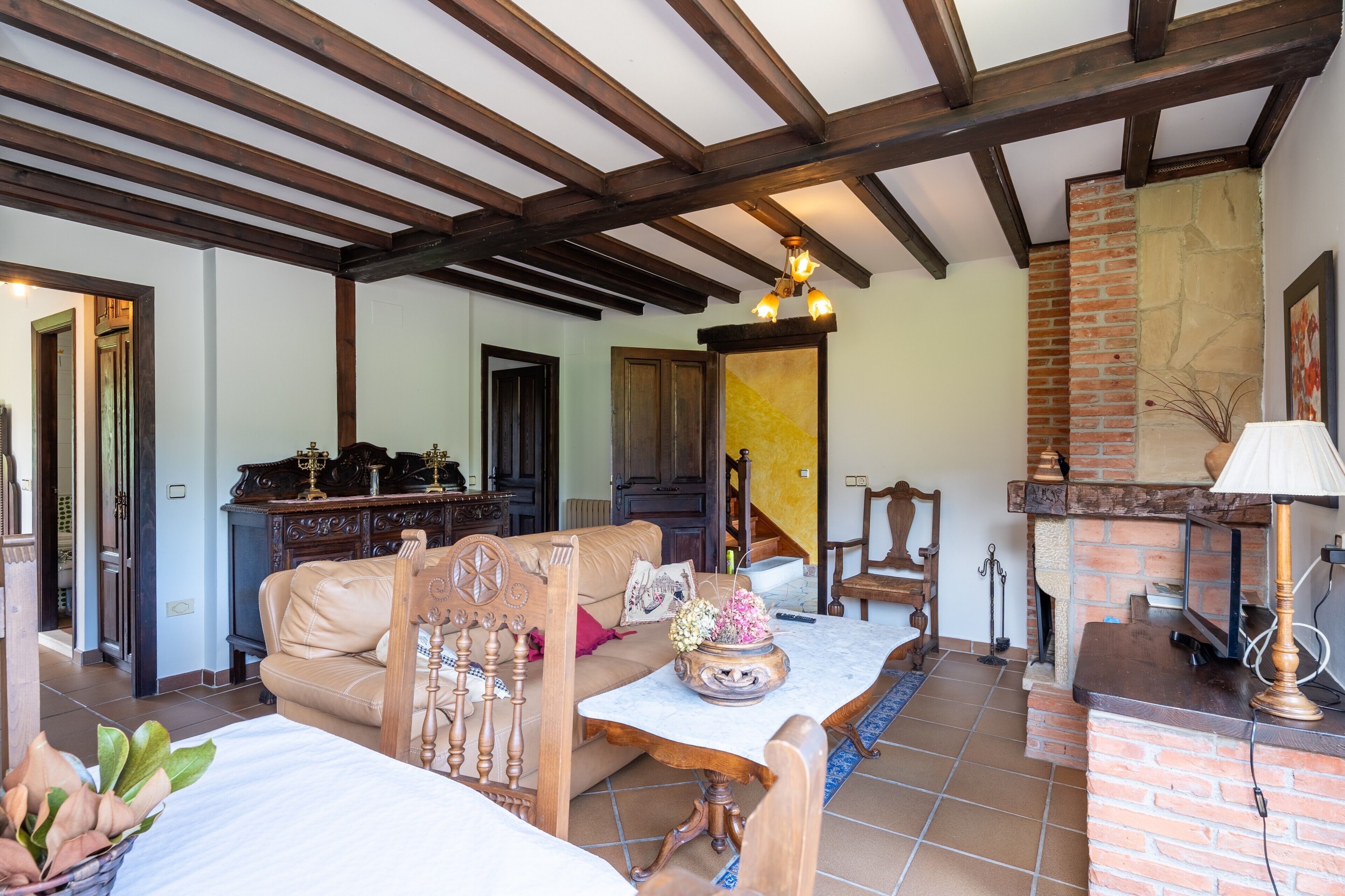 A room with a wooden ceiling, a fireplace, a sofa, a table with a vase, and a lamp.