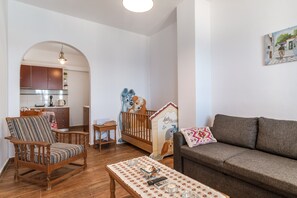 Una sala de estar con sofá, una silla de madera, una mesa con bandeja, una cuna y una cocina visible a través de un arco.