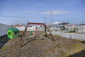 A playground with swings and slides, a small green house, and a fenced area with white chairs.