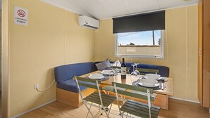 A compact room with a dining table set for two, a blue cushioned bench, a window with a view, and a ceiling-mounted air conditioning unit. - Holiday Home 'El Olivo 2' with Shared Pool, Wi-Fi & Air Conditioning (Chiclana de la Frontera)