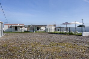 A row of mobile homes with a gravel driveway, a fenced area with lounge chairs, and a clear sky.