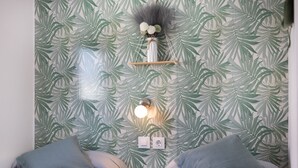 Un dormitorio con papel tapiz de hojas tropicales, una mesita de noche con un florero y dos almohadas.