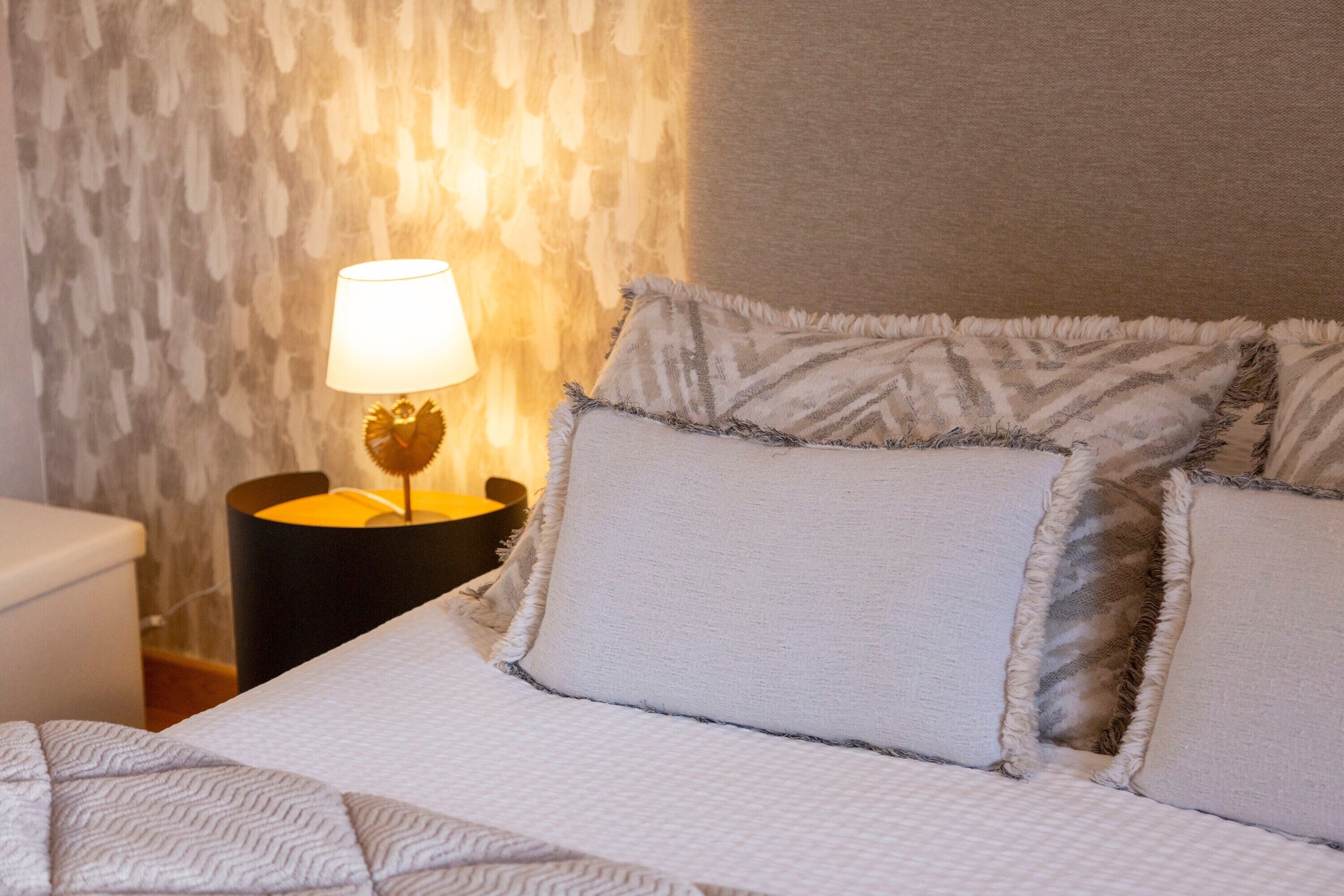 Ein ordentlich bezogenes Bett mit gemusterten Betttextilien, einer Nachttischlampe und einer strukturierten Wand dahinter.