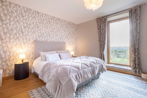Un dormitorio con cama, mesita de noche, lámpara, ventana con cortinas y vistas al exterior con vegetación.