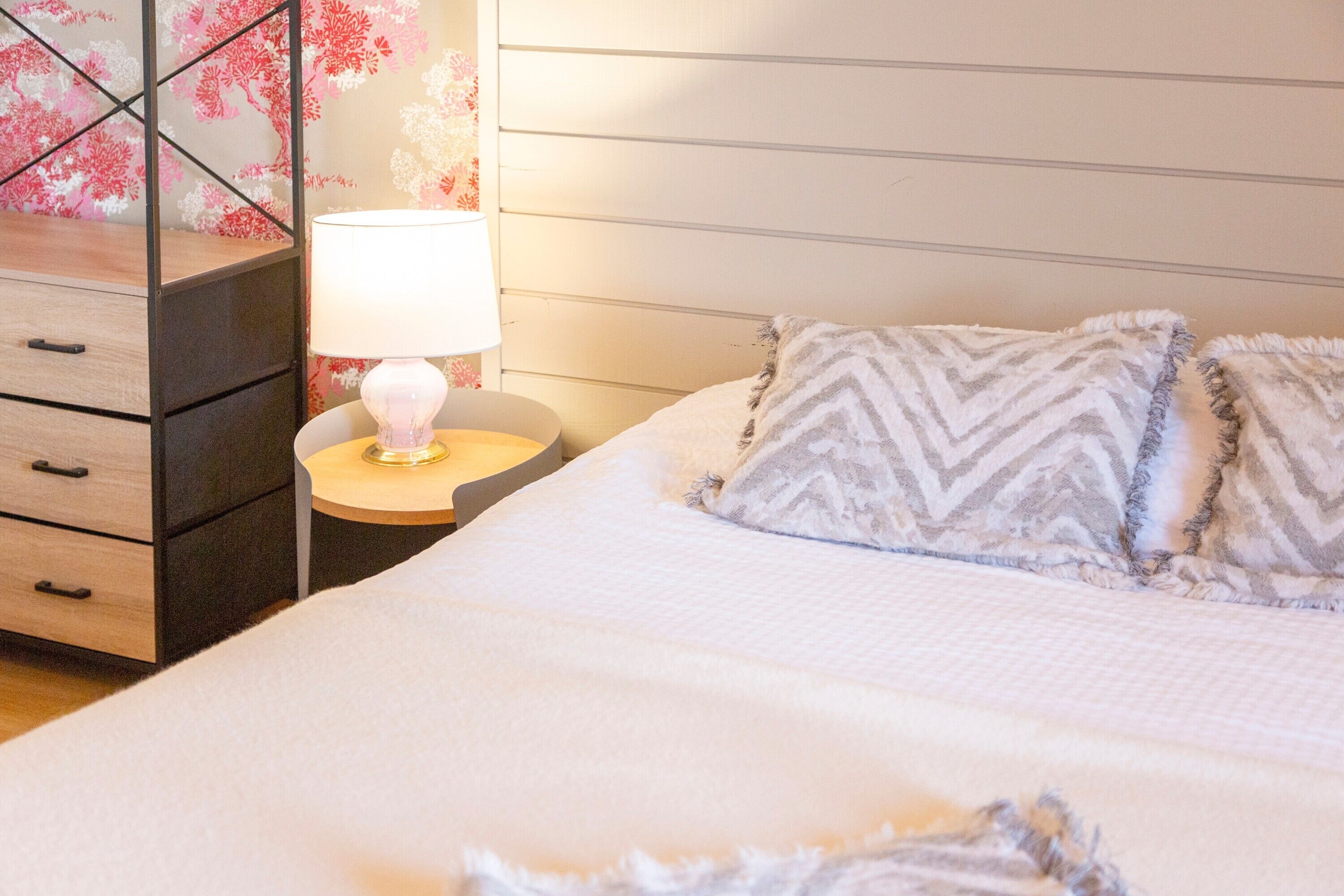 Une chambre à coucher avec un lit, une table de chevet avec une lampe, une commode et un mur recouvert d’un papier peint à motifs floraux.