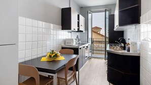 Una cocina moderna con azulejos blancos, una mesa de comedor con sillas, una pequeña isla con un lavamanos y vistas a un edificio a través de una puerta abierta.