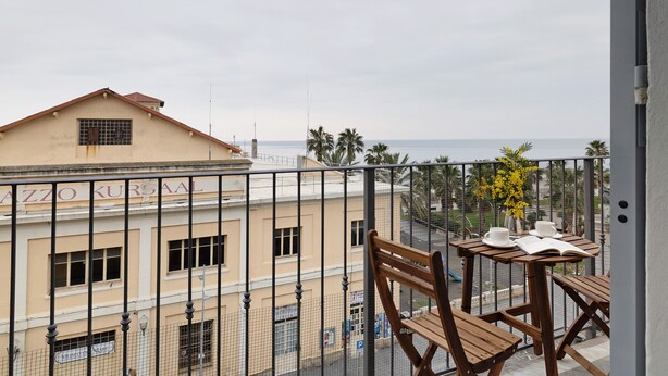 Un balcón con mobiliario de madera, vista al mar y un edificio con el letrero 'ZAZZO KURBAAL'.