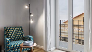 Una habitación con un sillón estampado, una pequeña mesa con un libro, una lámpara de pie y vistas a un balcón exterior a través de un ventanal.