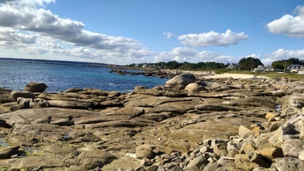 Un littoral rocheux parsemé de gros rochers, un ciel bleu dégagé et des maisons qui se dessinent à l’horizon.