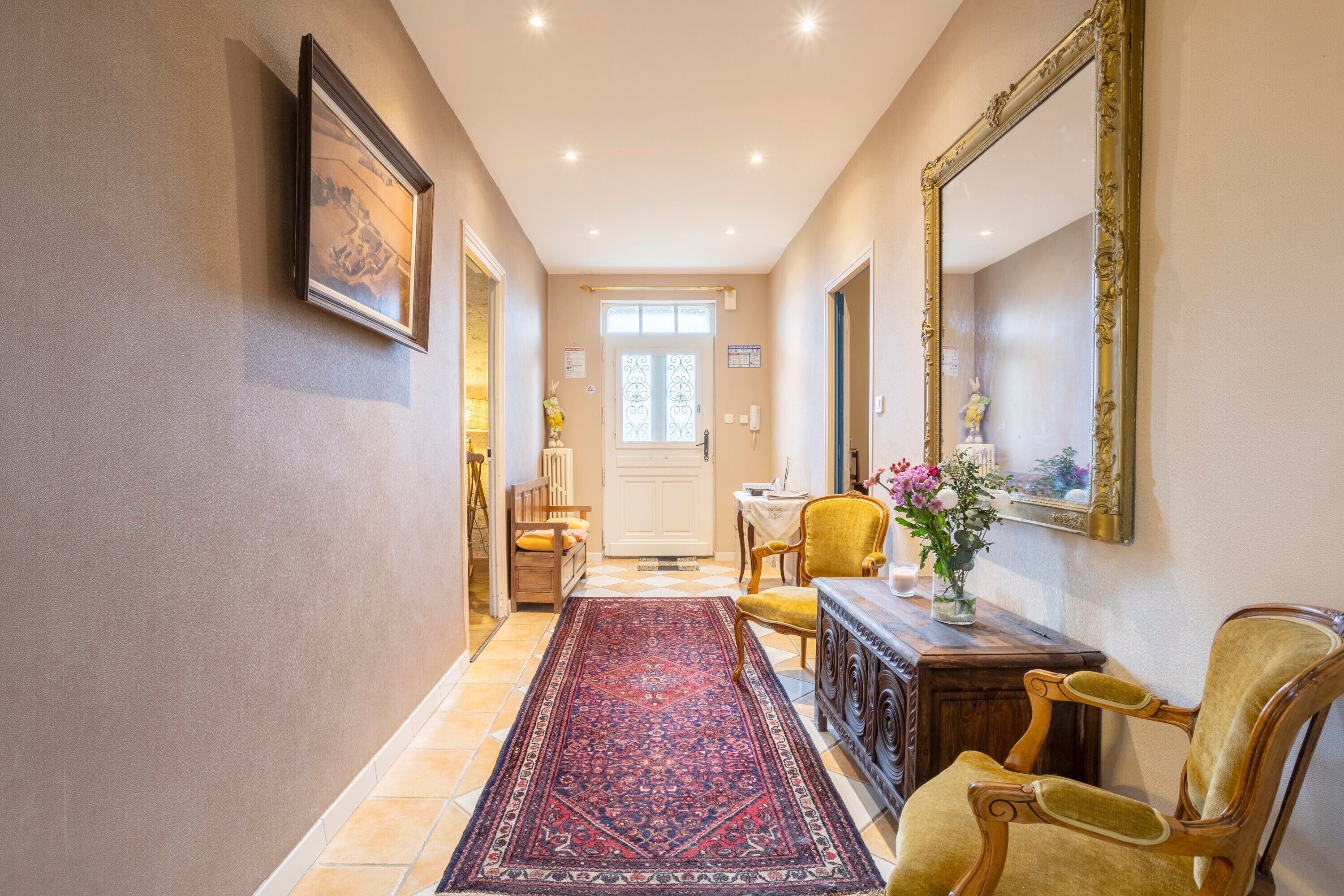 Un couloir avec un tapis à motifs, un banc en bois, une chaise jaune, une table avec un vase de fleurs et un grand miroir au mur.