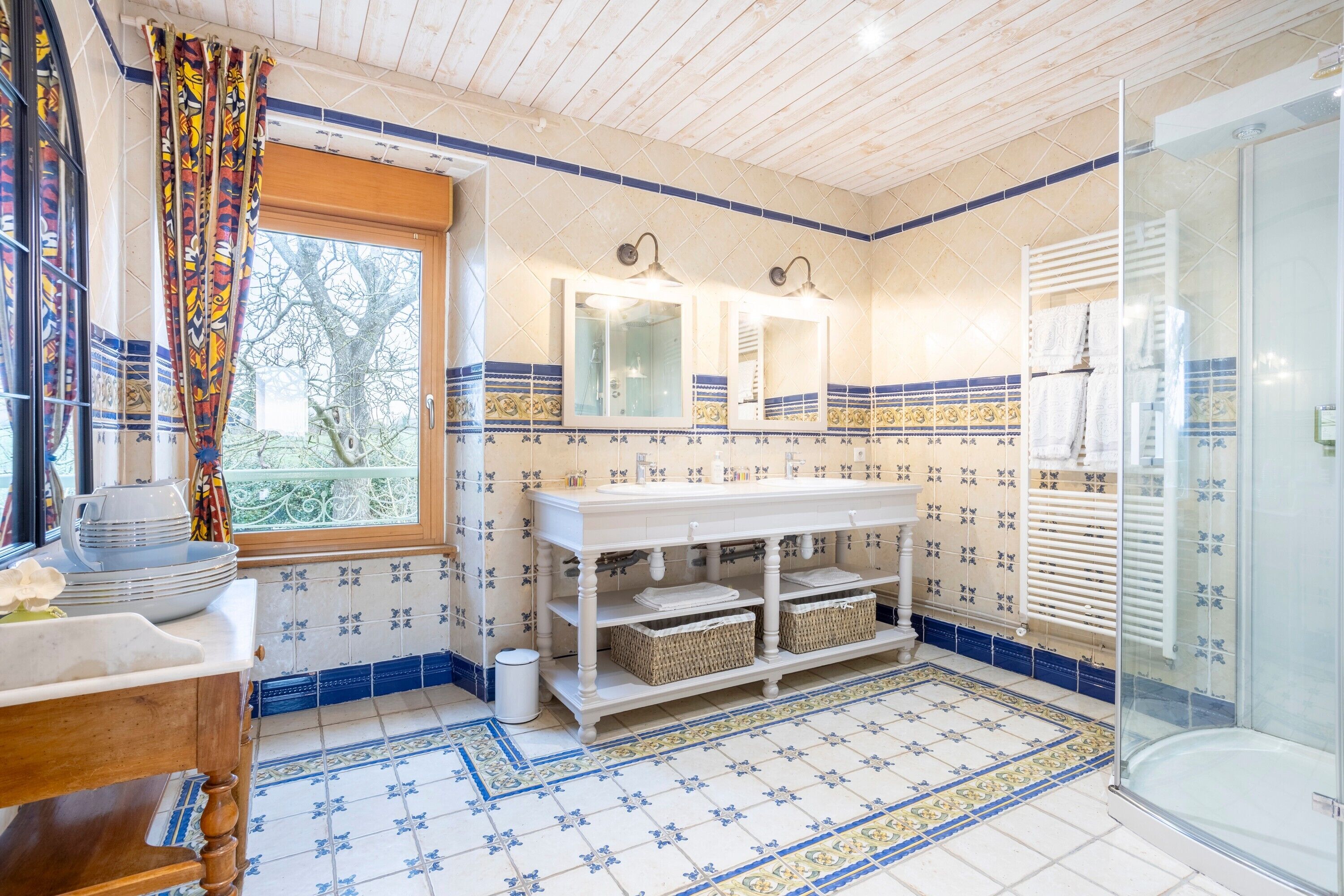 Een badkamer met een houten plafond, een raam met gordijnen, een wastafel met spiegel, een douche en betegelde vloeren.