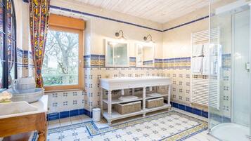 Een badkamer met een houten plafond, een raam met gordijnen, een wastafel met spiegel, een douche en betegelde vloeren.