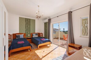 A bedroom with two beds, a chandelier, a balcony with a chair, and a view of the outside. - Holiday Home 'Breeze - Adults Only' with Sea View, Wi-Fi and Air Conditioning (Albufeira)