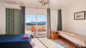Una habitación con cama, una mesita, una silla, un sofá, una araña y un cuadro en la pared, además de vistas a través de una puerta de cristal.