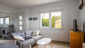 Ein modernes Wohnzimmer mit einem grauen Sofa, einem weißen Couchtisch und einem Fernseher auf einem Holzständer. Im Hintergrund ist eine Waschmaschine zu sehen.