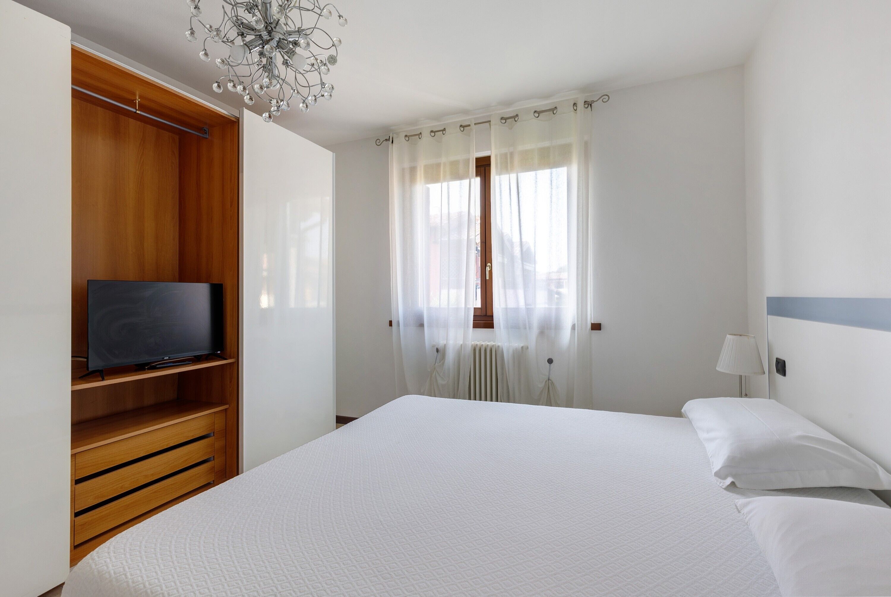A bedroom with a bed, a TV on a wooden shelf, a window with sheer curtains, and a bedside lamp.