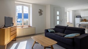 A modern living room with a sofa, a coffee table, a TV on a wooden cabinet, and a view through a large window.