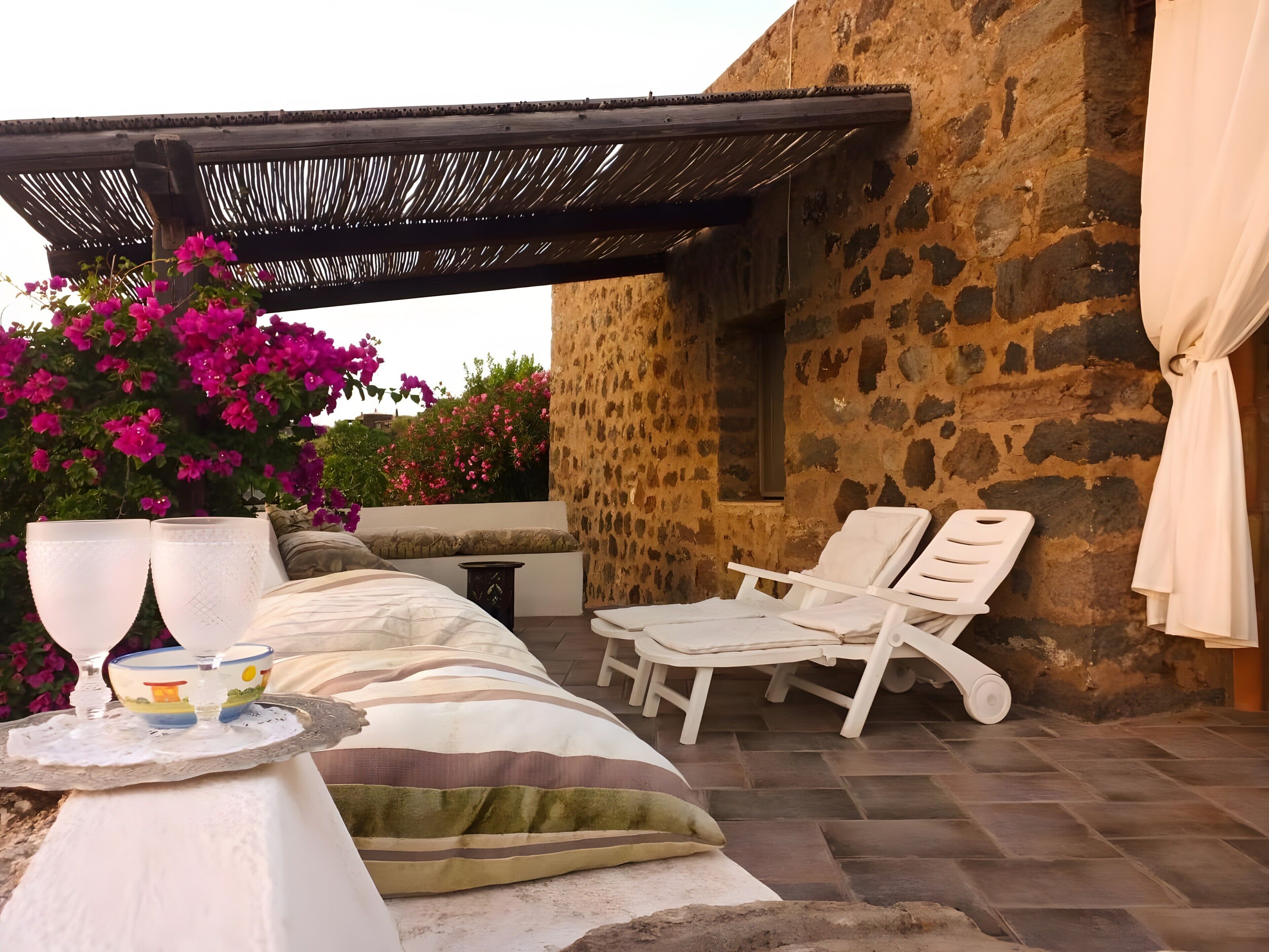 Ein Steinhaus mit überdachter Außenbestuhlung, einem Tisch mit Gläsern und einer Schale, und rosa Bougainvillea-Blumen.