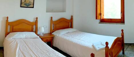 Dos camas de madera con ropa de cama blanca, una mesita de noche con lámpara, un cuadro enmarcado en la pared y una ventana con marco de madera.