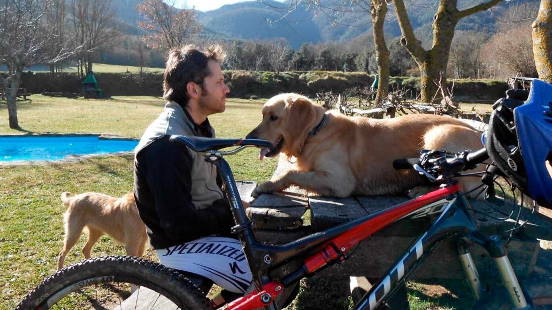 Una persona sentada en una bicicleta con un perro a su lado, en un parque con una piscina y árboles.