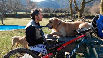 Una persona sentada en una bicicleta con un perro a su lado, en un parque con una piscina y árboles.