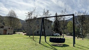 Un espacio al aire libre con césped, un conjunto de columpios, un edificio de piedra y árboles.