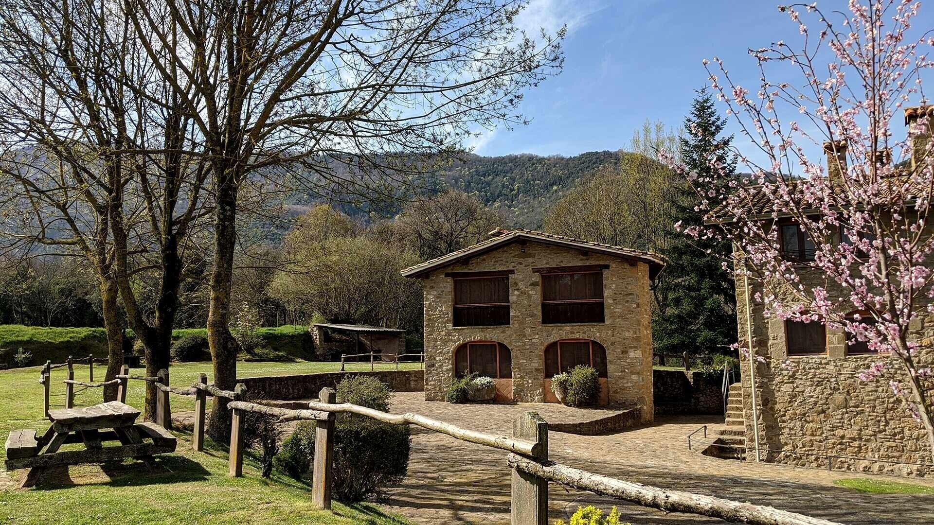 Un edificio de piedra con techo inclinado, rodeado por una cerca de madera, situado en un paisaje rural con árboles y colinas al fondo.