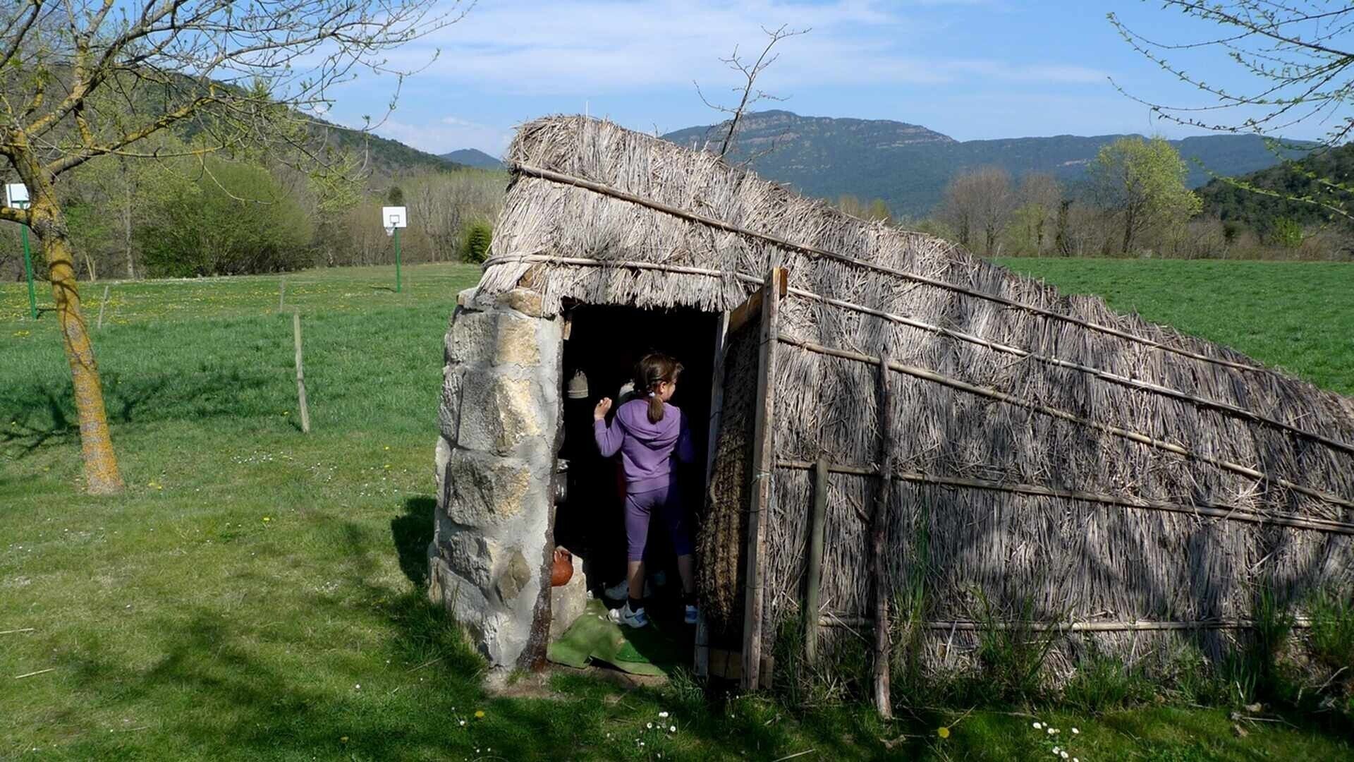 En rustik stenbyggnad med ett halmtak, en basketkorg och en gräsyta, med berg i bakgrunden.