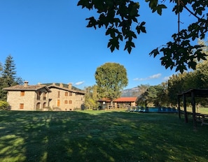 A stone building with a red roof, a large tree, and a wooden structure in a grassy area.