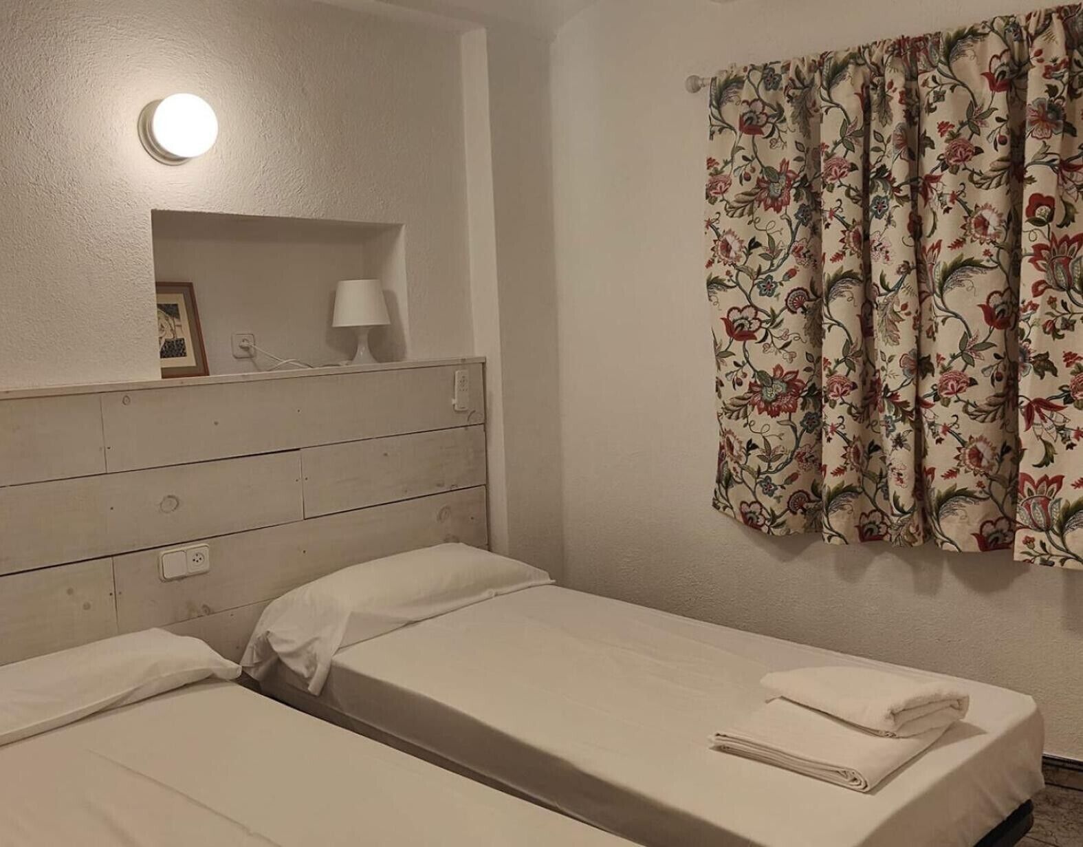 Ein Zimmer mit zwei Betten, einem Nachttisch mit Lampe, einem blumigen Vorhang und einer Wandleuchte.