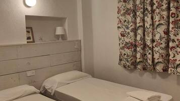 Ein Zimmer mit zwei Betten, einem Nachttisch mit Lampe, einem blumigen Vorhang und einer Wandleuchte.